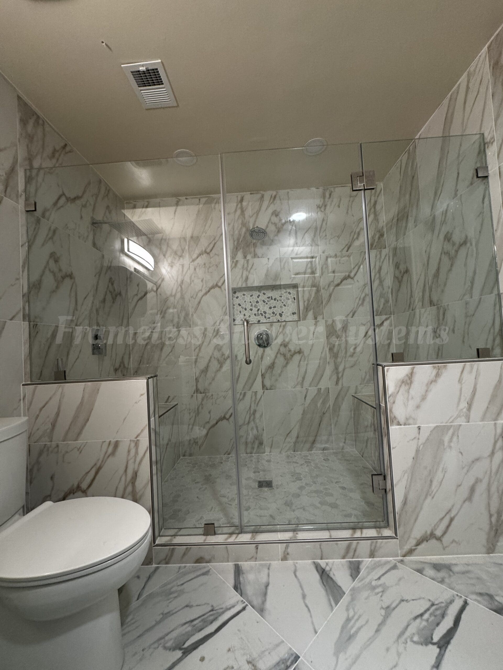 Frameless glass shower enclosure with chrome hardware, marble tiled walls, and a pebble mosaic niche