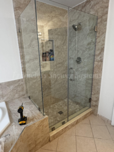 Frameless glass shower enclosure with beige marble tile and a cordless drill on the adjacent ledge