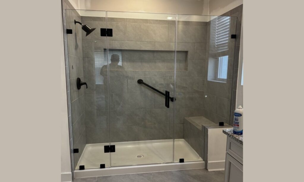 Walk-in shower with gray tiled walls, frameless glass door, matte black fixtures, built-in bench, and a grab bar