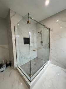 Walk-in glass shower enclosure with brushed brass hardware, white marble tile walls, and a mosaic floor