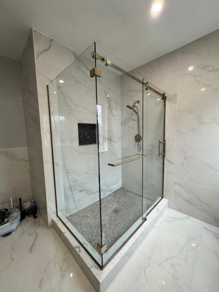 Walk-in glass shower enclosure with brushed brass hardware, white marble tile walls, and a mosaic floor