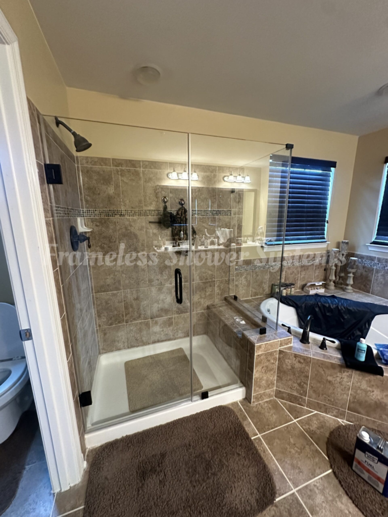 Frameless glass shower enclosure with dark bronze hardware in a tan tiled bathroom next to a soaking tub